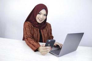 une jeune femme islamique asiatique portant un foulard sourit sur un téléphone portable avec un ordinateur portable sur la table. photo