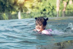 petit garçon asiatique joue dans l'eau tout en nageant dans la piscine portant des brassards. photo