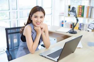 belle femme asiatique professionnelle est assise sur une chaise pour travailler en souriant joyeusement au bureau, elle regarde la caméra qui a un ordinateur portable et une lampe posée sur la table. photo