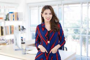 belle femme asiatique professionnelle portant une chemise à manches longues dame de bureau debout et souriant au travail souriant joyeusement au bureau elle regarde la caméra et met sa main dans sa poche. photo