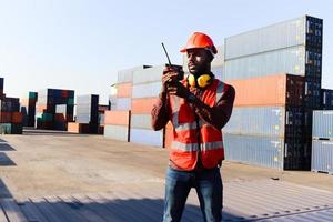 portrait d'un jeune travailleur ingénieur afro-américain portant un gilet et un casque de sécurité de couleur rouge néon vif, parlant à un collègue avec talkie-walkie dans la cour de conteneurs de fret d'expédition logistique. photo