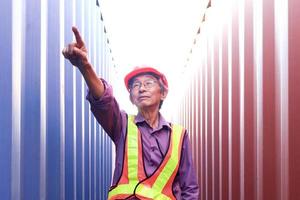 portrait d'un ingénieur senior asiatique âgé portant un gilet de sécurité et un casque, pointant vers la caméra, debout entre l'arrière-plan des conteneurs rouges et bleus dans la cour des conteneurs de fret d'expédition logistique. photo