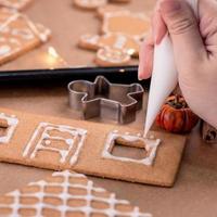 la femme décore la maison des biscuits au pain d'épice avec une garniture de crème glacée au glaçage blanc sur fond de table en bois, du papier sulfurisé dans la cuisine, gros plan, macro. photo