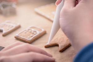 la femme décore la maison des biscuits au pain d'épice avec une garniture de crème glacée au glaçage blanc sur fond de table en bois, du papier sulfurisé dans la cuisine, gros plan, macro. photo