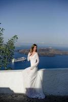 belle fille blonde mince femme top modèle mariée aux cheveux longs et aux jambes dans la robe de bal de mariée blanche avec un long train et des volants se dresse sur une île pittoresque de santorin en grèce photo