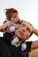 une petite fille mignonne est assise sur son cou près de son père. père et fille marchent dans le champ photo