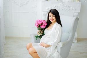 photo en gros plan d'une femme enceinte avec un ventre nu tenant un bébé chaussons blancs et fleurs roses sur un lit blanc