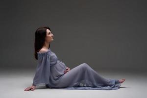 une jeune femme enceinte vêtue d'une robe se dresse sur un fond gris. photo