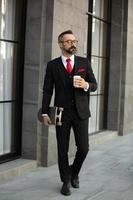 homme d'affaires hipster en costume tenant une planche à roulettes avec une tasse de café près du bureau en plein air