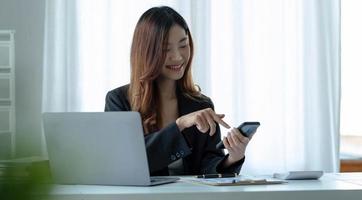 portrait d'une jeune femme d'affaires asiatique a utilisé un smartphone pour écrire un ordinateur portable dans le bureau à domicile. belle fille à l'ordinateur de bureau emporter la commande à la maison. start-up asie femme télémarketing PME en ligne. photo