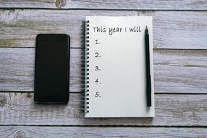 cette année, je vais envoyer un texte sur un bloc-notes sur un fond en bois avec un smartphone et un stylo. photo