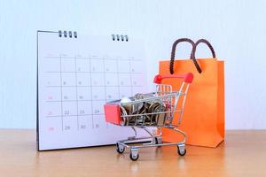 calendrier avec jours et panier supermarché et sac en papier orange sur table en bois. notion de magasinage photo