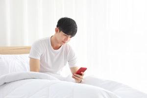 homme asiatique portant un tissu blanc utilisant un téléphone portable pour vérifier les nouvelles du matin et les médias sociaux juste après s'être réveillé sur son lit avec espace de copie photo