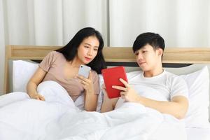 couple asiatique se regardant téléphone tout en s'allongeant dans le lit en planifiant leur prochain mariage photo