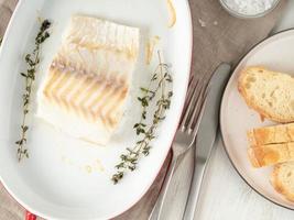 filet de morue, fraîchement cuit au four avec du thym dans un plat en porcelaine pour la cuisson, pain sur une assiette, vue de dessus photo