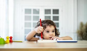 jolie petite fille assise au bureau à la maison pour faire ses devoirs, lire, écrire et peindre. les enfants peignent. les enfants dessinent. enfant d'âge préscolaire avec des livres à la maison. les enfants d'âge préscolaire apprennent à écrire et à lire. bambin créatif photo