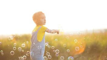 une fille en combinaison denim souffle des bulles de savon en été dans un champ au coucher du soleil. journée internationale des enfants, enfant heureux, activités de plein air. fond d'été. mode de vie sain et respectueux de l'environnement photo