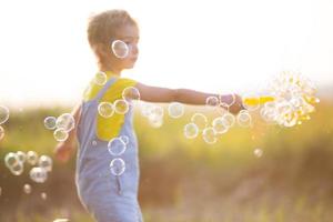 une fille en combinaison denim souffle des bulles de savon en été dans un champ au coucher du soleil. journée internationale des enfants, enfant heureux, activités de plein air. fond d'été. mode de vie sain et respectueux de l'environnement photo