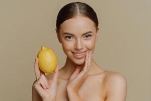 photo horizontale d'une femme européenne assez saine utilise du citron pour fabriquer un masque facial naturel prend soin de son corps et de sa peau se dresse seins nus à l'intérieur isolé sur fond marron. notion de beauté.