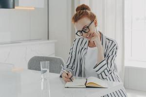 une femme européenne prend des notes dans un planificateur personnel sourit doucement tient des poses de stylo sur le bureau photo