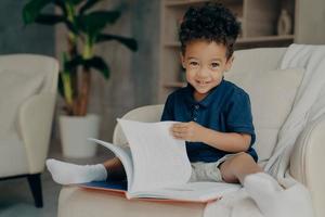 mignon petit garçon afro-américain assis dans un fauteuil avec un livre d'histoire photo