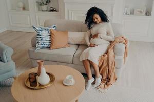 belle photo femme afro-américaine assise sur un canapé confortable et attendant bébé, regardant vers le bas sur son ventre