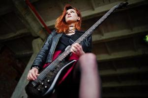 fille punk aux cheveux rouges portant une jupe noire et rouge, avec une guitare basse à un endroit abandonné. portrait de musicienne gothique. photo