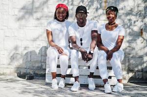 trois amis afro-américains élégants, portent des vêtements blancs assis sur un banc. mode de rue des jeunes noirs. homme noir avec deux filles africaines. photo