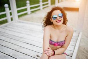 portrait d'une jolie fille posant en bikini avec des lunettes de soleil debout sur une terrasse en bois blanche.