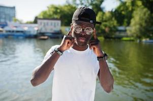 garçon afro-américain élégant, porter des vêtements blancs, des lunettes et une casquette contre la jetée du lac. mode de rue des jeunes noirs. photo