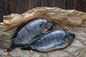 Deux tilapias entiers non cuits sur du papier ciré et fond de bois sombre à plat photo