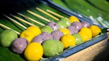 bonbons à base de farine verte, violette, jaune, brochettes posées sur des feuilles de bananier vertes. photo