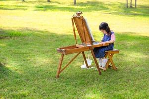 une petite fille est assise sur le banc en bois et peinte sur la toile posée sur un pupitre à dessin photo