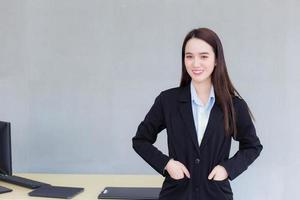 une femme asiatique travaillant dans une entreprise professionnelle se tient debout et met sa main dans la poche d'un costume noir pendant qu'elle sourit joyeusement au bureau en arrière-plan. photo