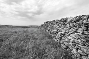 une photo en niveaux de gris d'un champ avec un mur sous un ciel nuageux