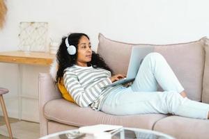 femme afro-américaine dans un casque discutant en ligne sur un ordinateur portable en position allongée sur un canapé dans le salon photo