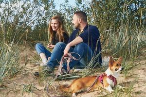 jeune couple heureux d'homme et de femme avec un chien corgi assis sur le sable. deux personnes avec animal de compagnie photo