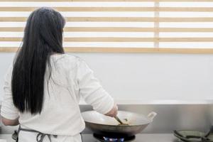 une femme asiatique aux longs cheveux noirs cuisine des aliments dans une casserole en acier sur une cuisinière avec un espace de copie derrière. photo