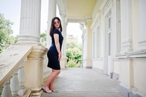 fille brune en robe noire, lunettes de soleil posées contre la vieille maison vintage, dans la rue de la ville. photo