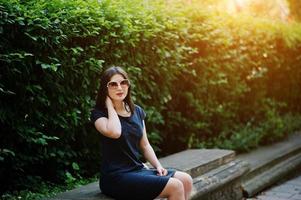 fille d'affaires brune en robe noire sur des lunettes de soleil assise à la frontière contre des buissons, posant dans la rue de la ville. photo