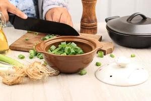 échalote d'oignon vert tranché sur un bol brun, trancher une main asiatique féminine sur fond à l'aide d'un couteau noir. processus de cuisson dans la cuisine photo