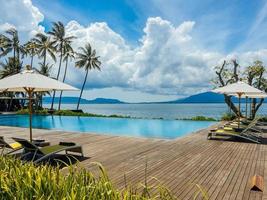 belle piscine tropicale dans un hôtel ou un complexe avec parasol, chaises longues en cocotier, palmiers avec vue sur la piscine à débordement, fond océan et montagne.