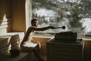 jeune homme versant de l'eau sur des pierres chaudes dans le sauna photo