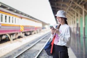 une belle femme asiatique tenant un sac et un smartphone à la recherche d'informations de voyage. en attendant le train sur le quai photo