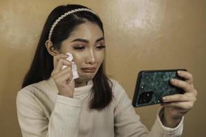 triste adolescente qui pleure en vérifiant son téléphone assis. femme asiatique regardant l'écran du smartphone versant des larmes photo