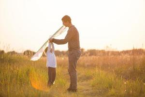 papa aide sa fille à faire voler un cerf-volant dans un champ en été au coucher du soleil. divertissement familial en plein air, fête des pères, fête des enfants. zones rurales, soutien, entraide. lumière orange du soleil