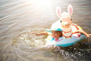 fille au chapeau nageant dans la rivière avec cercle gonflable en forme de lama. alpaga gonflable pour un enfant. mer au fond sablonneux. vacances à la plage, natation, bronzage, crèmes solaires.
