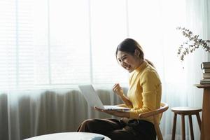 jeune femme avec ordinateur portable exprimant son enthousiasme dans le bureau à domicile, une femme asiatique excitée se sent euphorique en lisant de bonnes nouvelles en ligne. photo