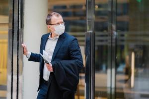 protection respiratoire, coronavirus, maladie dangereuse. un homme sérieux se tient dans les portes d'un immeuble de bureaux, porte un masque médical, tient un téléphone portable moderne, un journal pour lire un article sur le traitement covid-19 photo