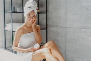 photo d'une jeune femme européenne souriante touche la joue, applique une crème hydratante sur le visage, a une expression heureuse, enveloppée dans une serviette de bain douce, pose dans la salle de bain. concept de routine d'hygiène beauté